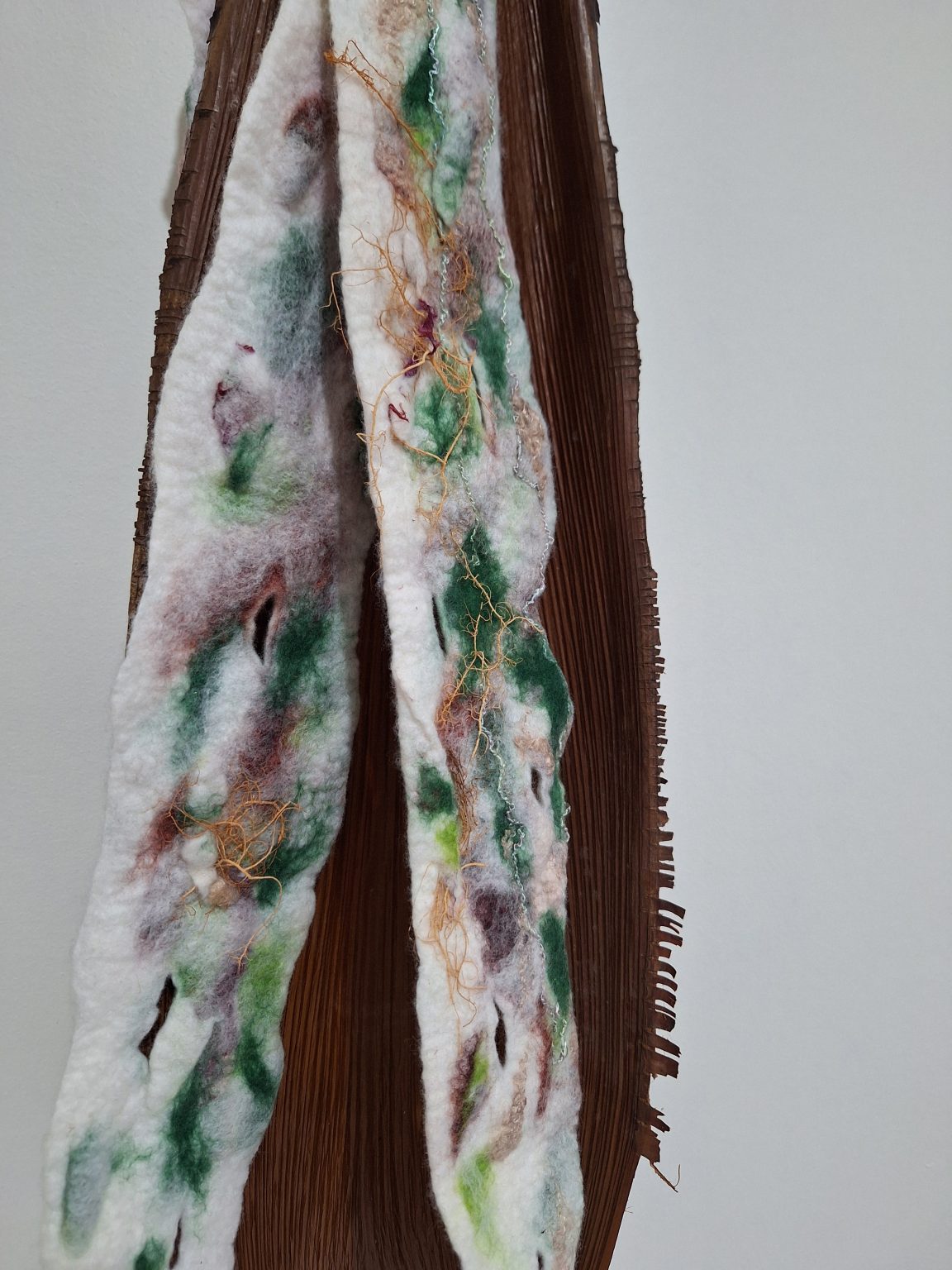Felted wool artwork with embroidery, dried lichen, and other threads hanging inside the base of a palm leaf. The contrast of natural world and the forms we meld together to create a new whole, together and separate.