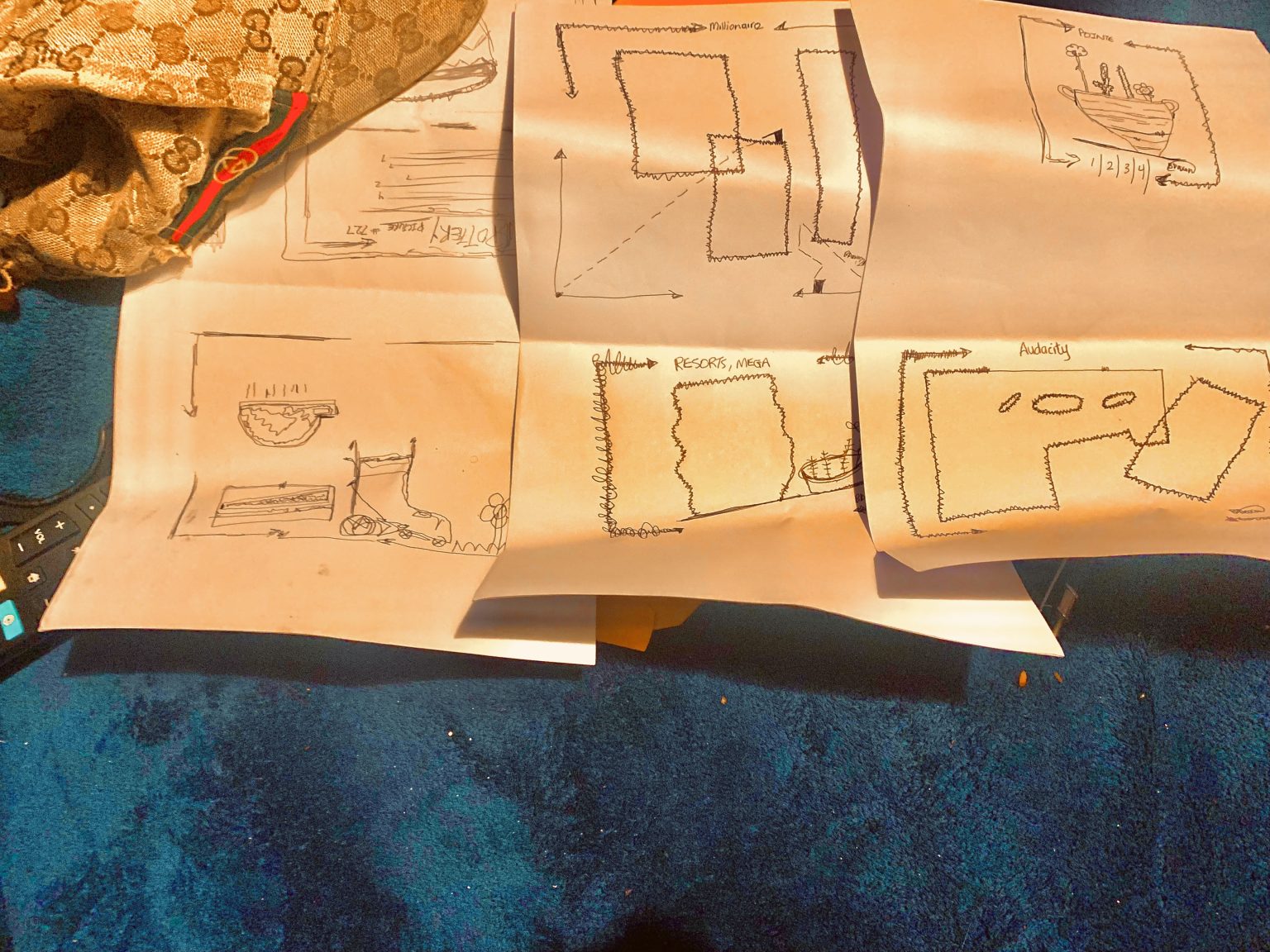 Hand-drawn architectural sketches spread across sheets of paper on a blue surface, each a rough plan annotated with a single word — "Millionaire", "Resorts, Mega", "Audacity", "Peasant" — pairing simple room layouts with small drawings of boats, beds and figures. Ambition and circumstance sketched as floor plans.