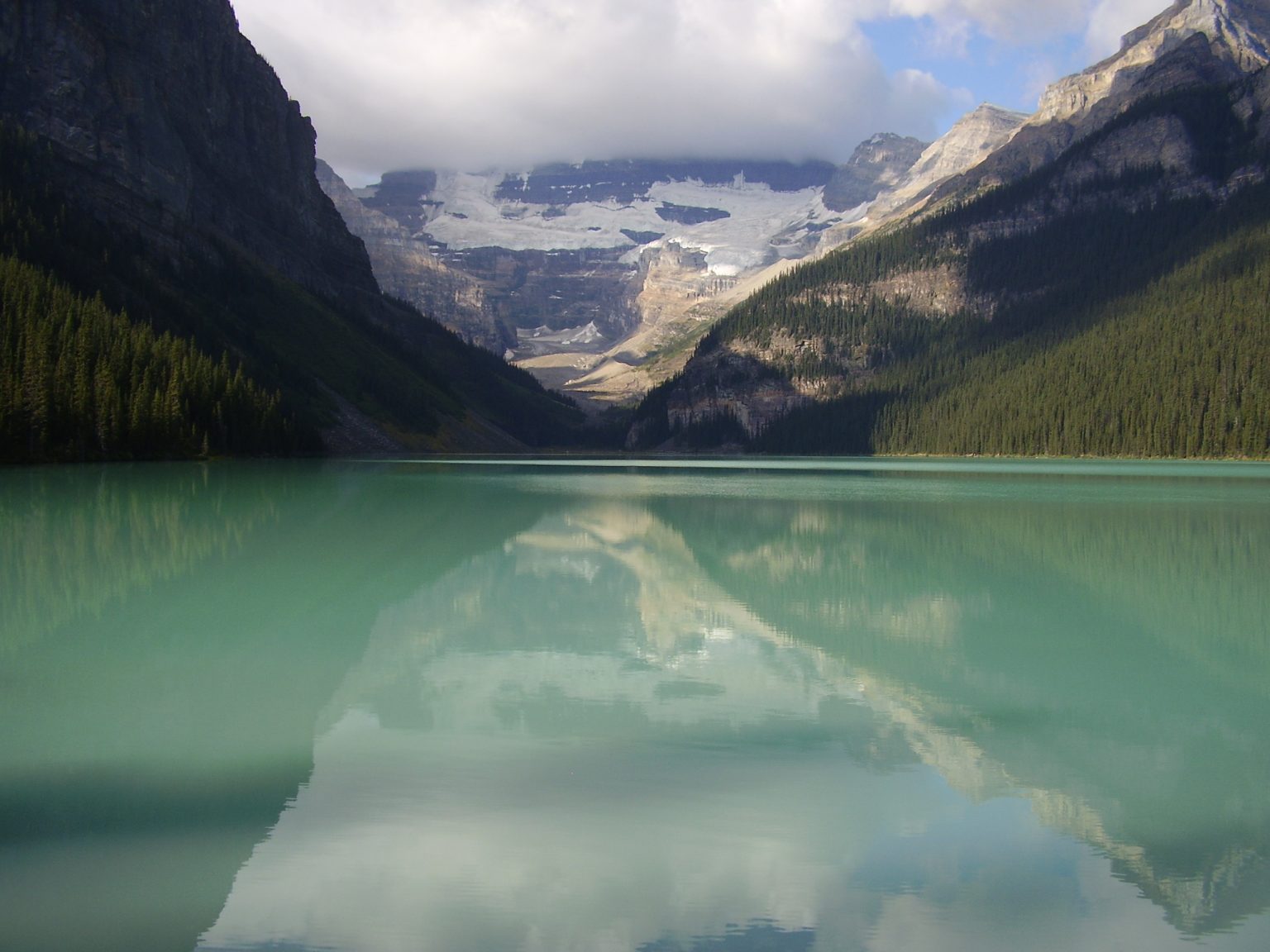 A photograph of Lake Louise in Banff, Alberta: turquoise glacial water holding the reflection of the peaks, dark spruce slopes closing in on both sides, and the Victoria Glacier under cloud at the head of the valley.